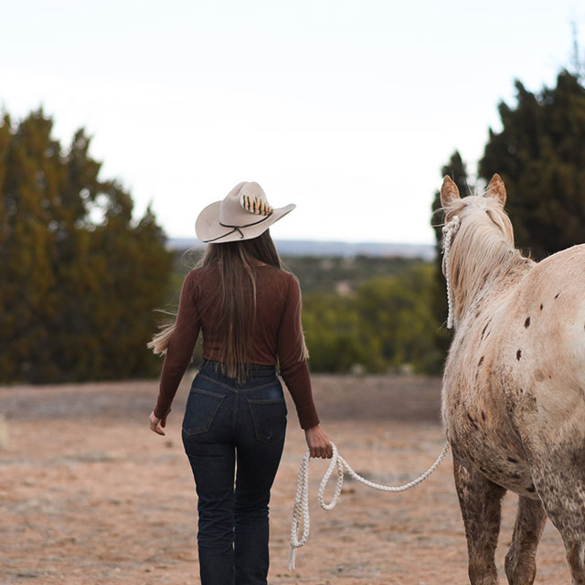 A woman, Stella Maria Baer, wearing a tan cowboy hat and leading an appaloosa horse away from the camera towards the trees.